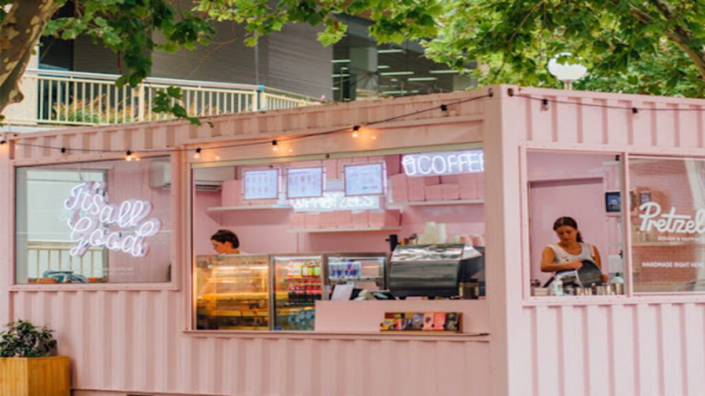 Charming pink shipping container converted into a coffee shop with neon 'It's all good' sign, serving window open, and employees at work, set in a vibrant outdoor setting.