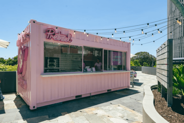 Outdoor pop-up food stand made from a pink shipping container labeled 'Pretzel', featuring a serving window and neon signs, set in a paved area with string lights overhead.