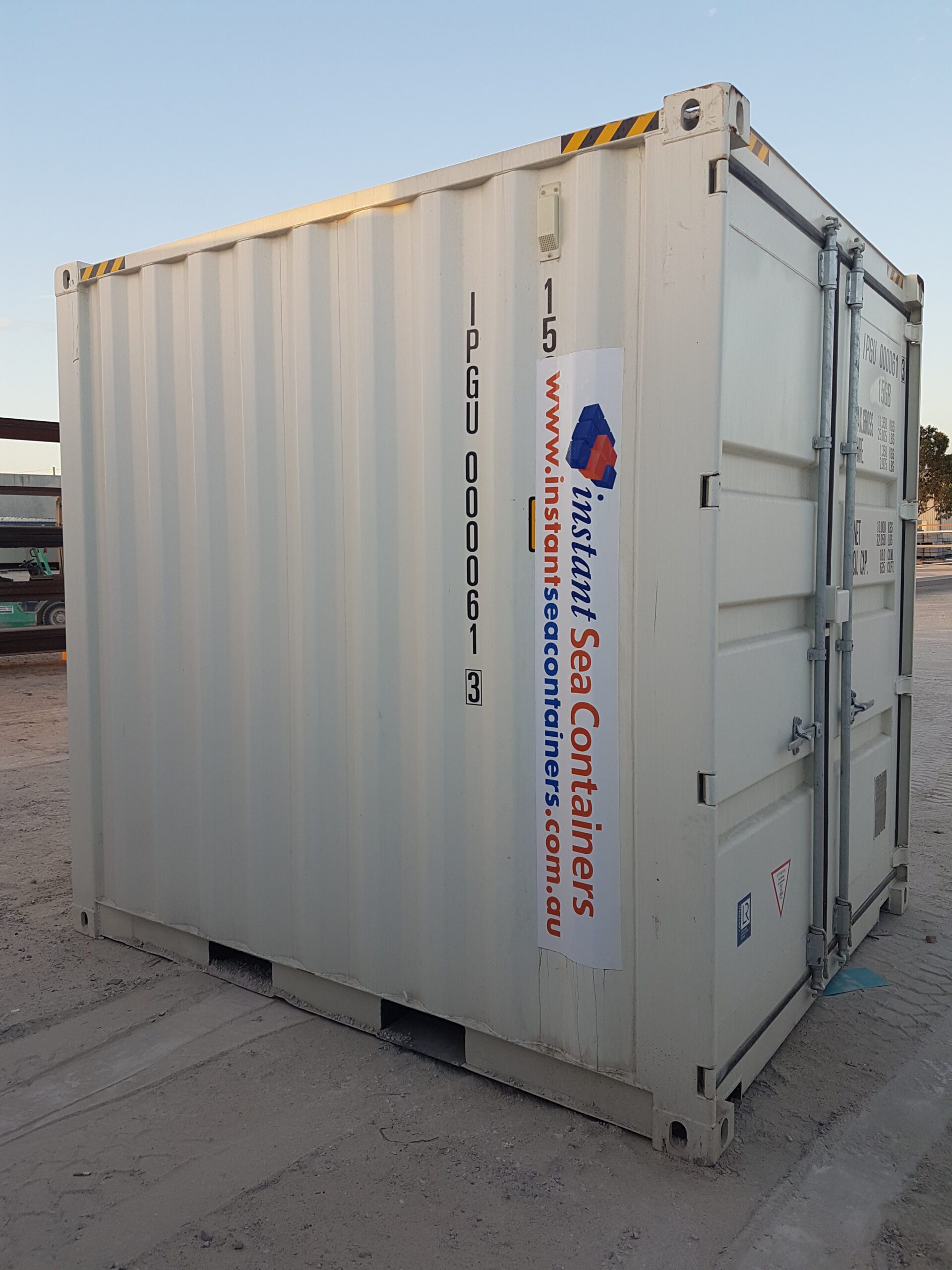White shipping container with company branding and safety stripes, positioned on a sandy ground during twilight.