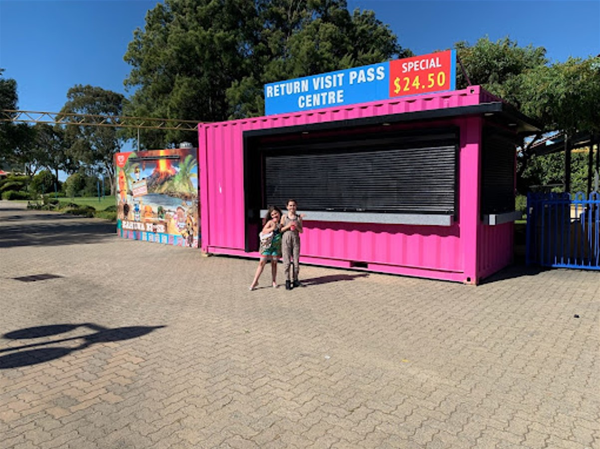 Outdoor ticket booth made from a repurposed pink shipping container labeled 'Return Visit Pass Centre' with a mother and child standing in front on a sunny day.