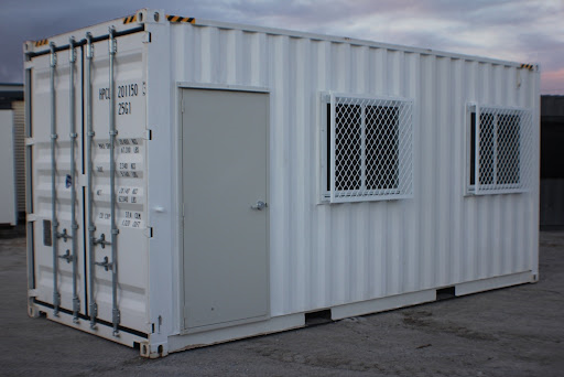 White shipping container converted into an office with a secure door and two windows with security bars, set in a yard during twilight.