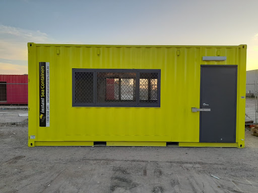Bright yellow shipping container modified into an office space, featuring a door and security windows, positioned on a gravel lot at dusk.