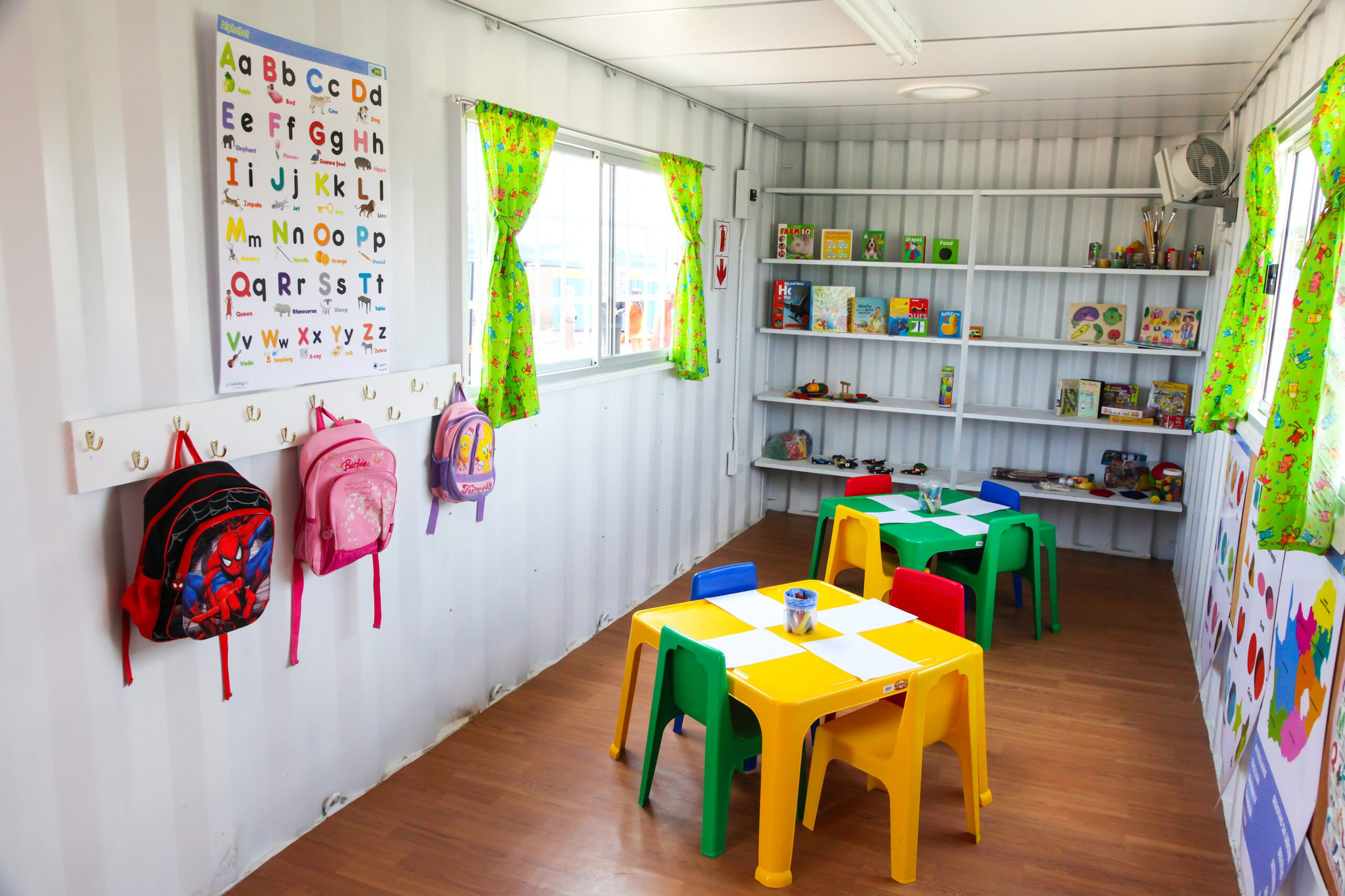 Colorful interior of a child's classroom inside a converted shipping container, featuring small tables, chairs, educational toys, and alphabet decorations on the walls.