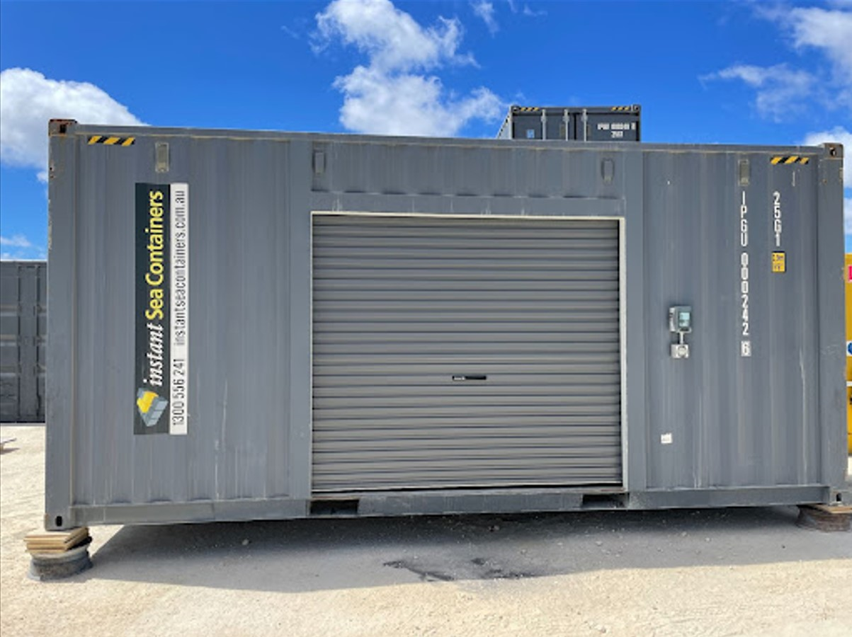 Gray shipping container modified with a large roll-up door and a side entrance, set on a gravel lot under a blue sky.