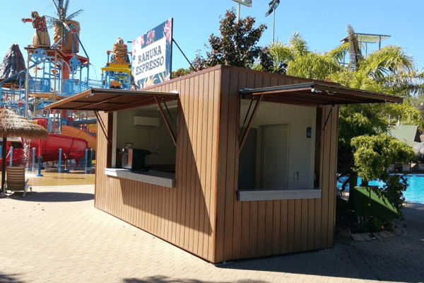 Outdoor café kiosk made from a shipping container with a wooden exterior, open service window, and a retractable awning, located beside a pool with waterpark attractions in the background.