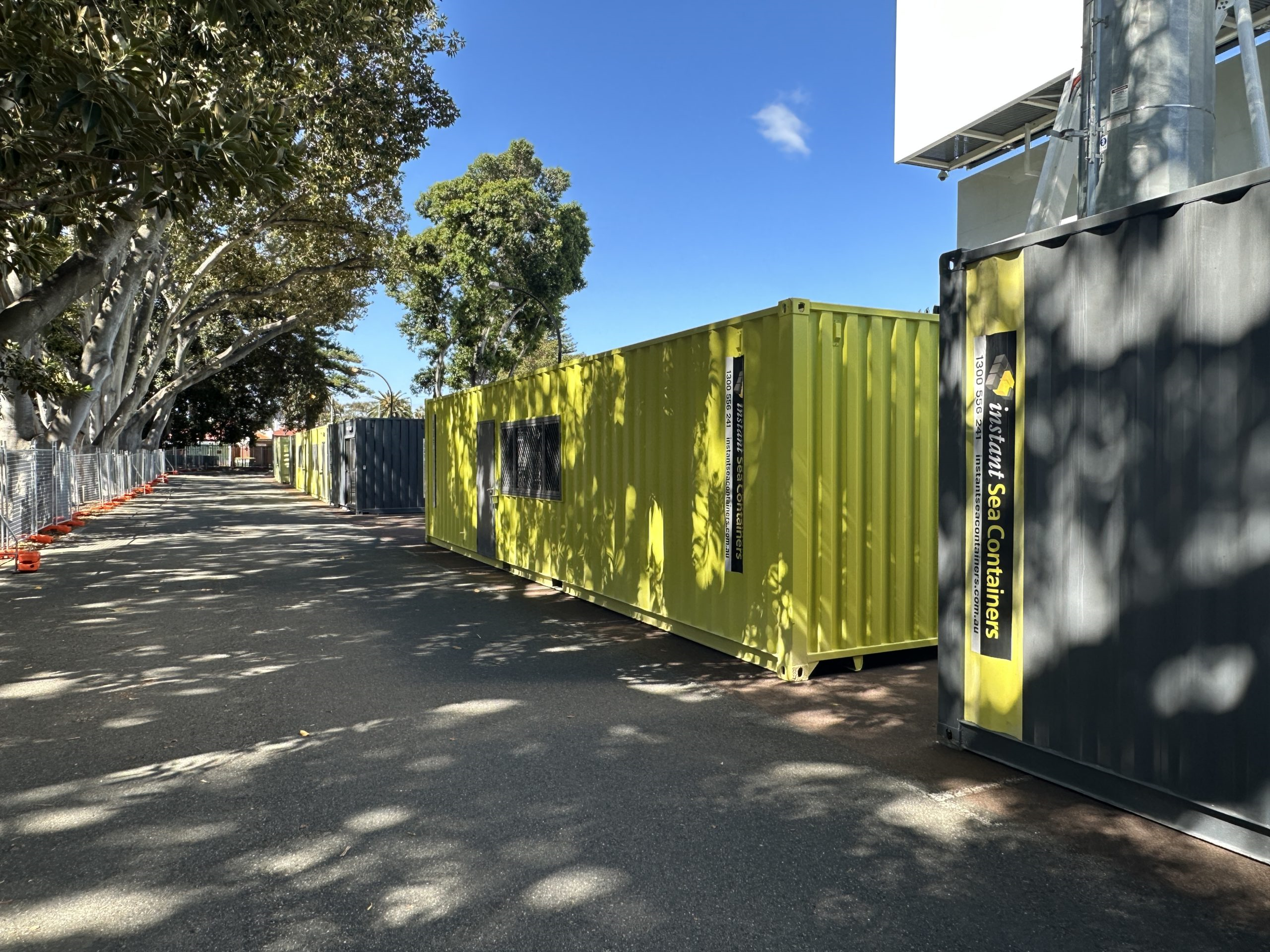 Yellow and gray shipping containers used as temporary offices, lined up along a shaded street with trees and dappled sunlight.