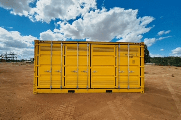 Bright yellow shipping container with multiple locking handles, placed on a dirt ground against a cloudy blue sky.