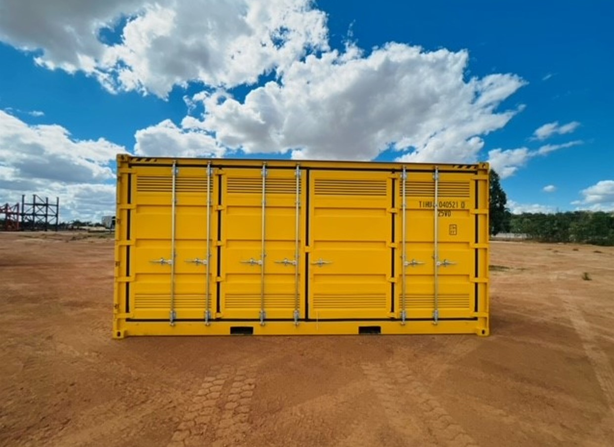 Bright yellow shipping container with multiple locking handles, placed on a dirt ground against a cloudy blue sky.