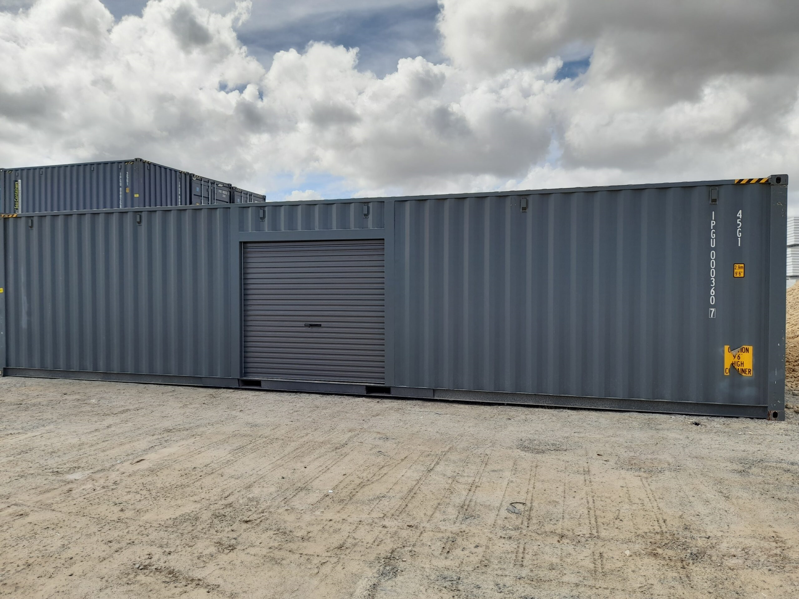 Large gray shipping container modified with a garage door, located on a gravel surface under a cloudy sky.