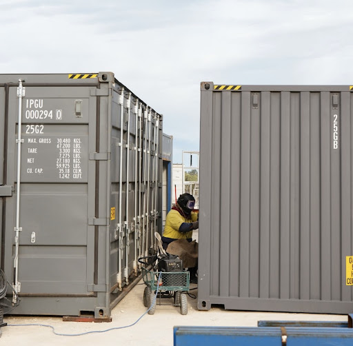 Worker welding between two gray shipping containers in an industrial outdoor setting.