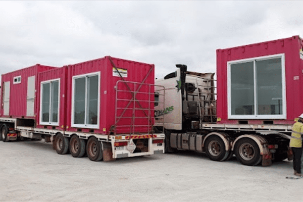Two modified red shipping containers on a flatbed truck, ready for transport, with a worker inspecting the setup.