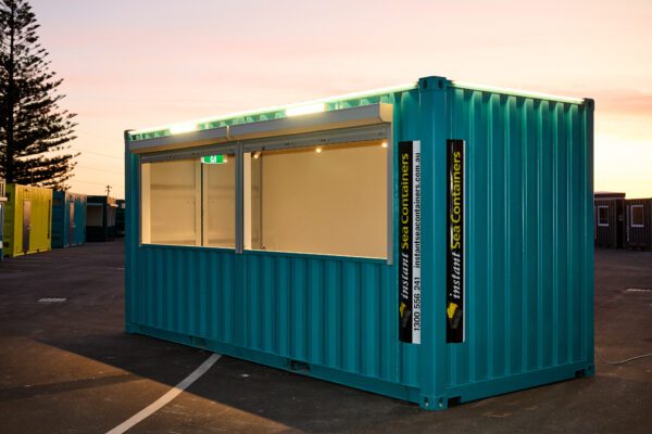 Teal shipping container modified for commercial use with large windows, illuminated under a sunset sky.