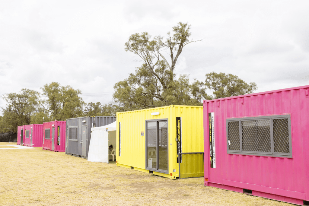 Colorful row of pink, yellow, and gray shipping containers repurposed as event offices in a grassy field with trees in the background.