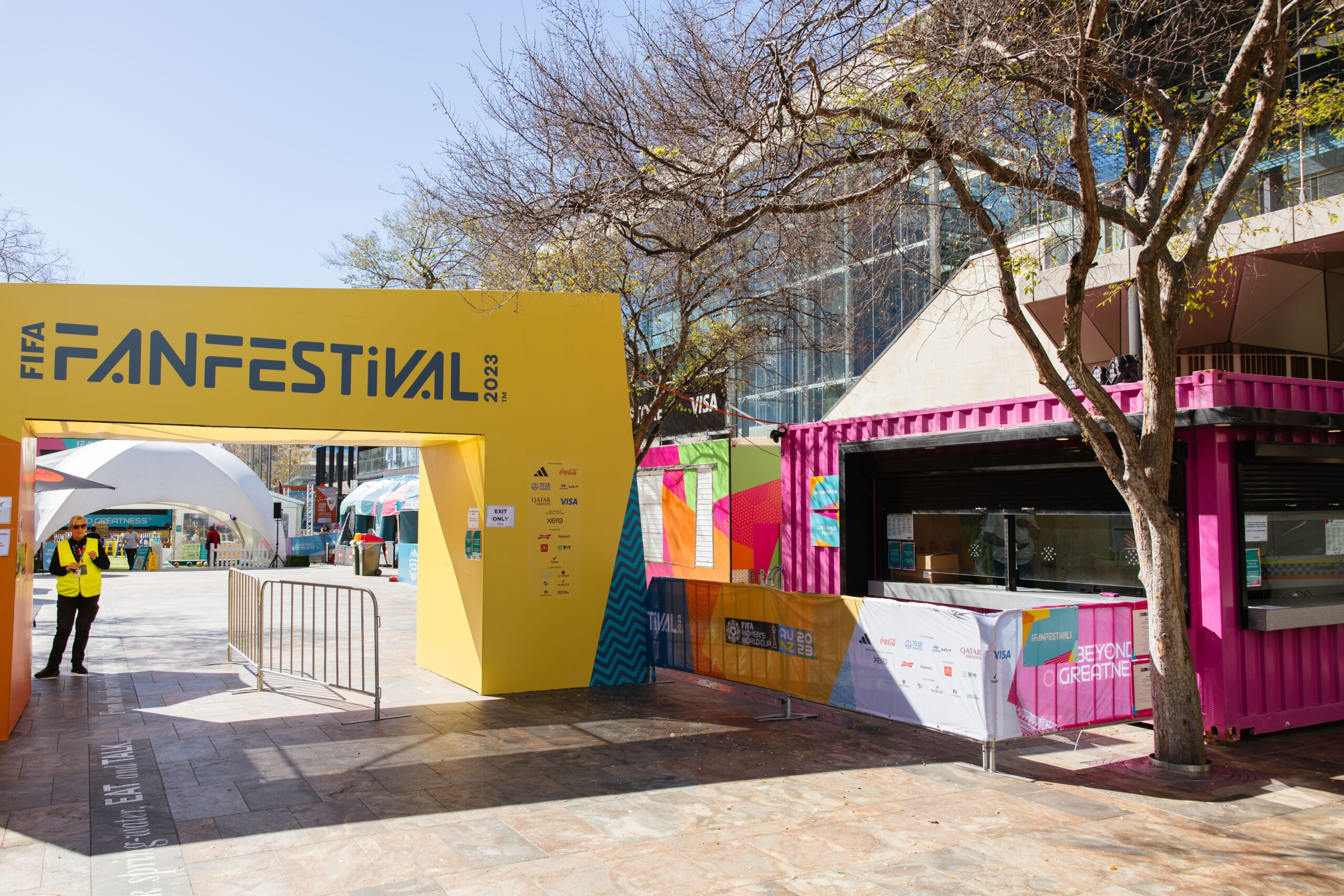 Entrance to the FIFA Fan Festival 2023 with colorful booths and trees in the background.