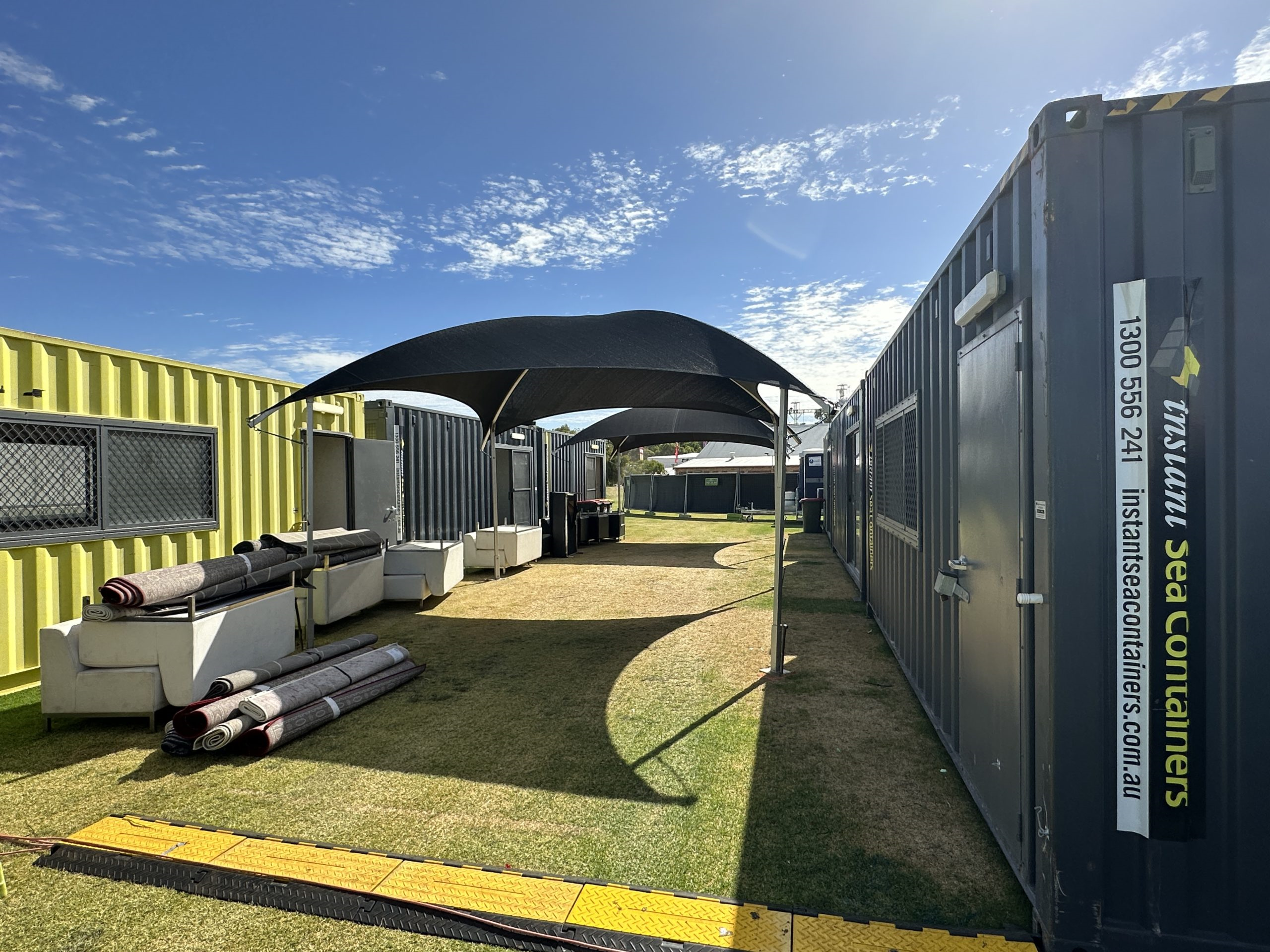 Outdoor event setup with modified shipping containers, artificial turf, and canopies, under a clear sky.