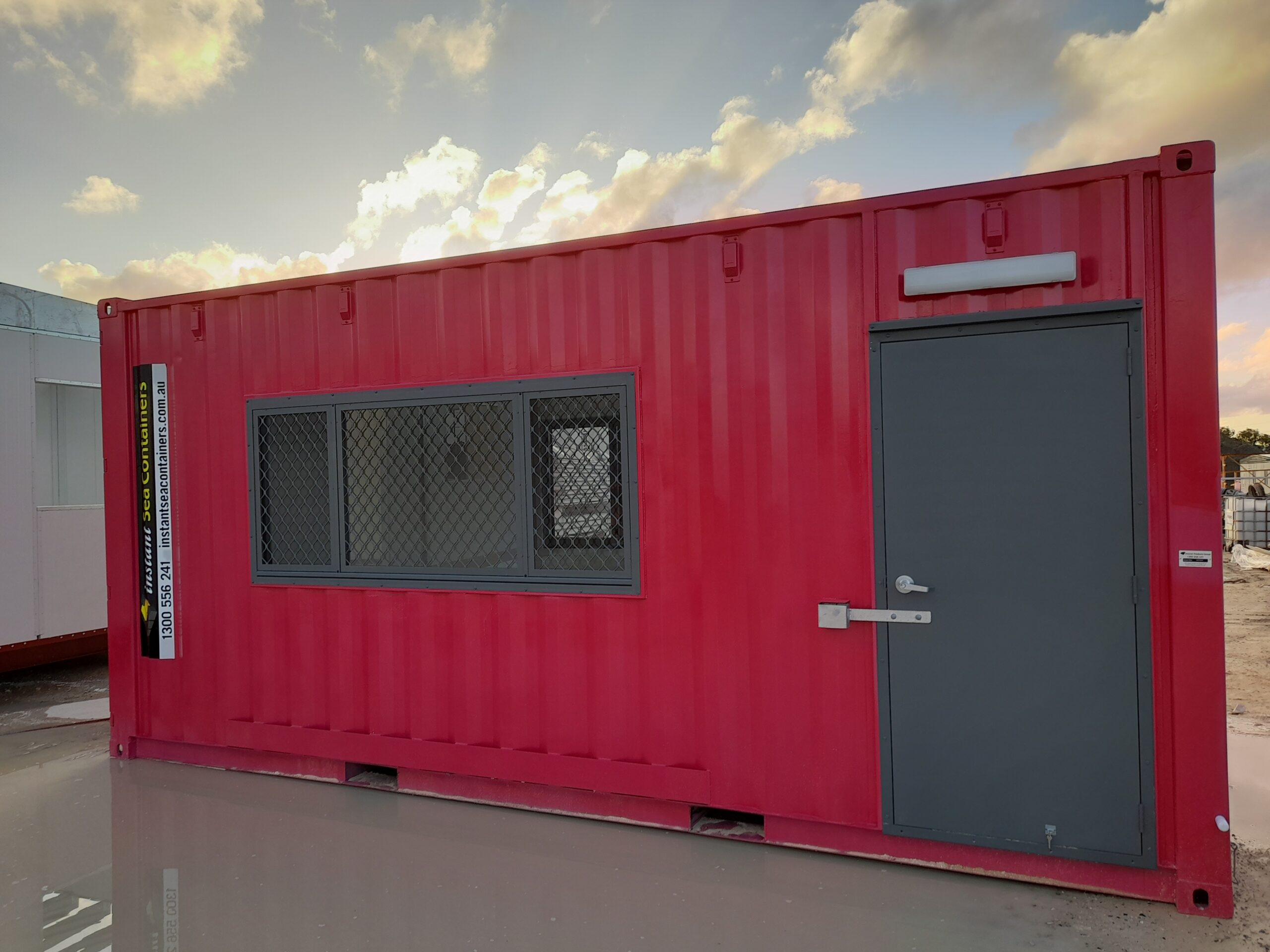 Red shipping container modified into an office with large security windows and a gray door, set against a cloudy sky background.