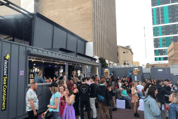 Crowd of people at an outdoor event featuring bars made from modified shipping containers in an urban setting.