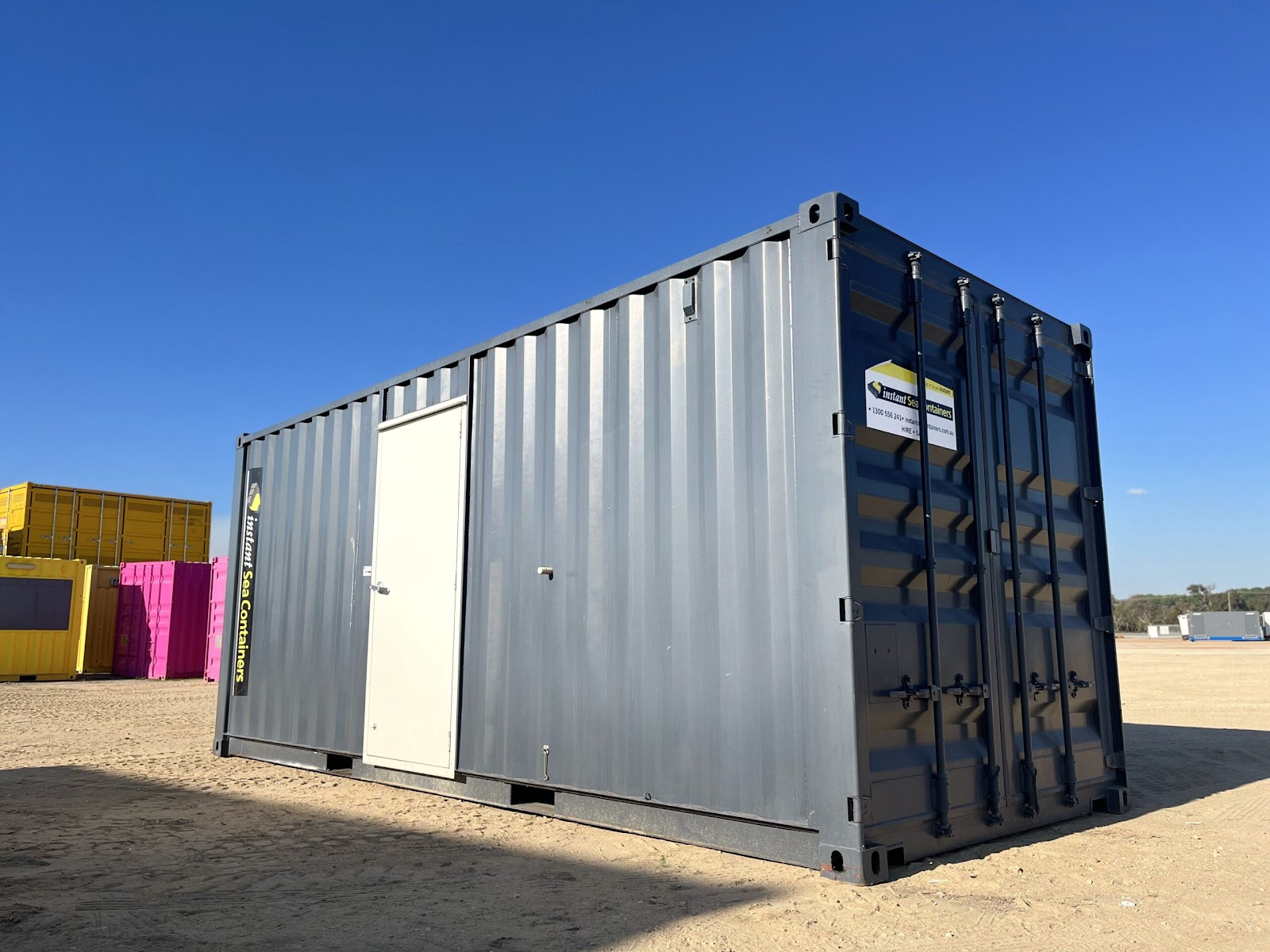 Gray shipping container with a white door, situated in an open lot with colorful containers in the background.
