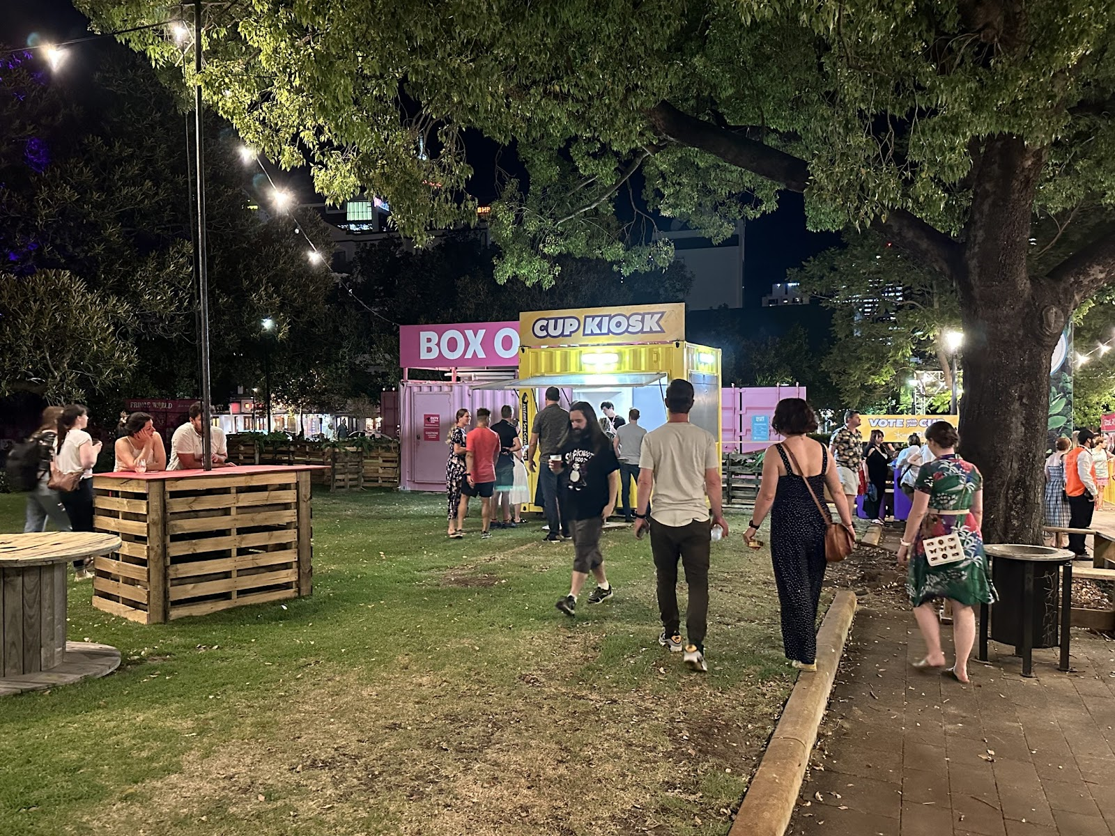 Nighttime scene at an outdoor event with people walking past colorful shipping container booths under tree canopy.