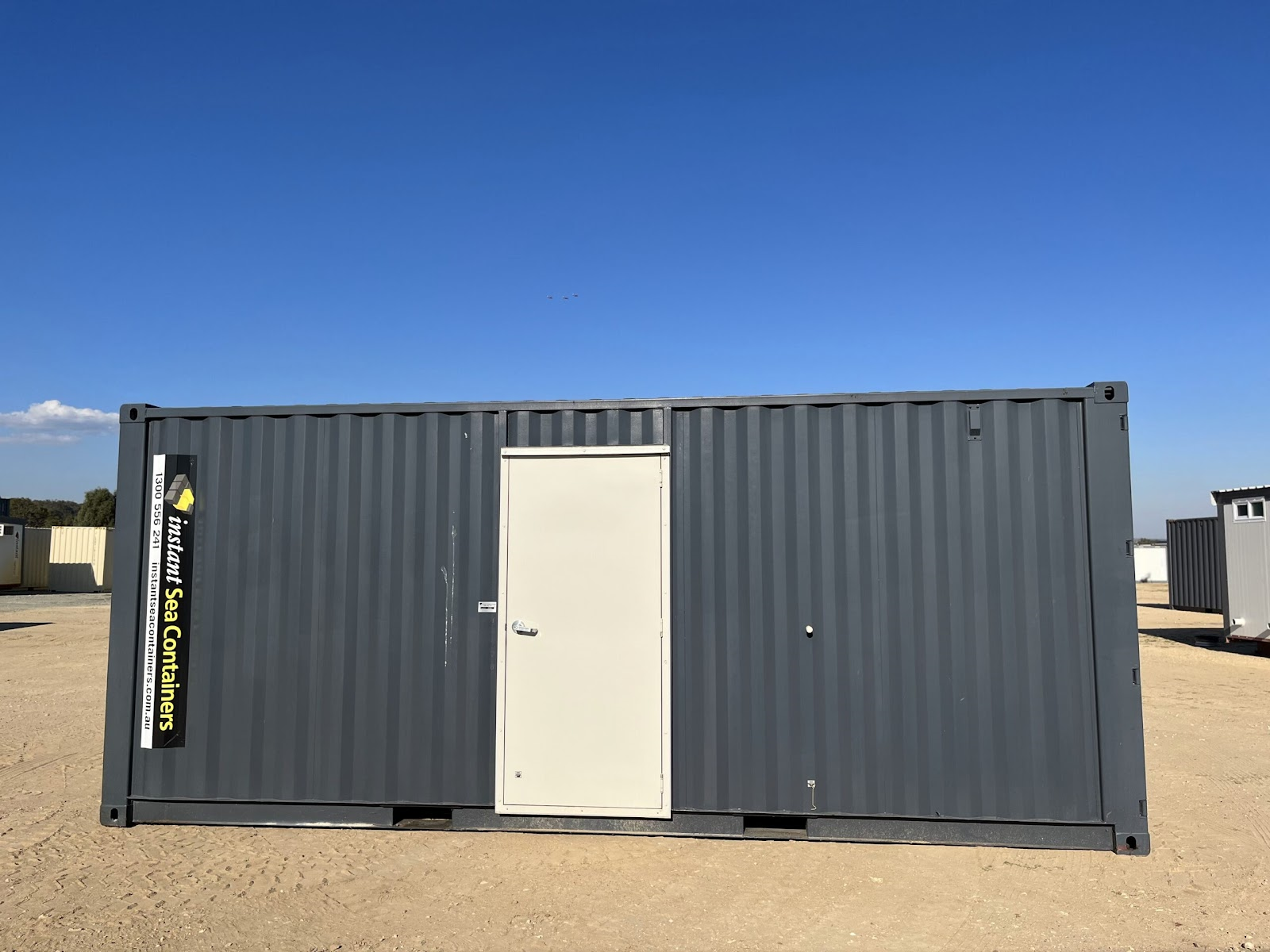 a standard gray shipping container fitted with a white door, placed in an open storage or construction area.