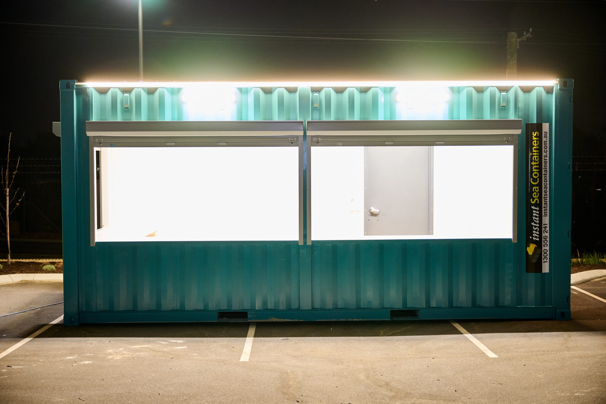 a teal shipping container at night, equipped with large display windows and external lighting.