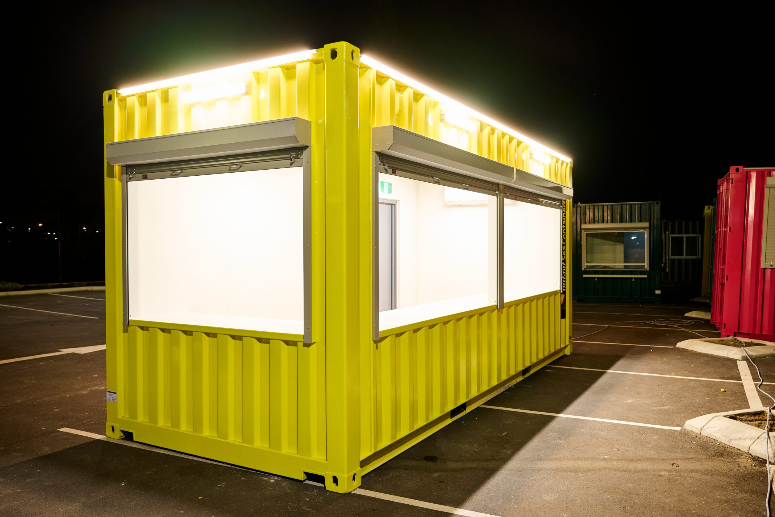 brightly lit, yellow Shipping Container Kiosk , given its large display windows and robust lighting setup for night-time operation