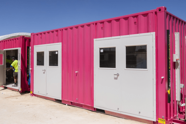 Bright pink shipping container converted into a modular building with multiple doors and windows, being worked on by a person wearing high-visibility clothing.