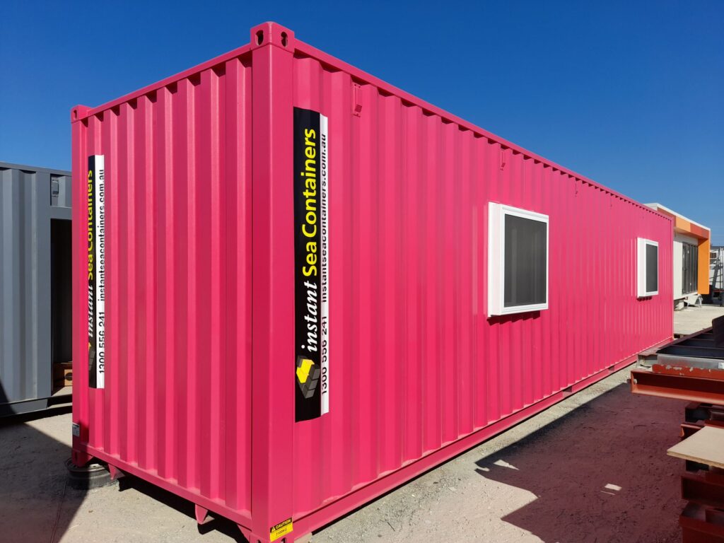 Bright pink shipping container office with two windows and Instant Sea Containers branding, positioned outdoors on a sunny day.