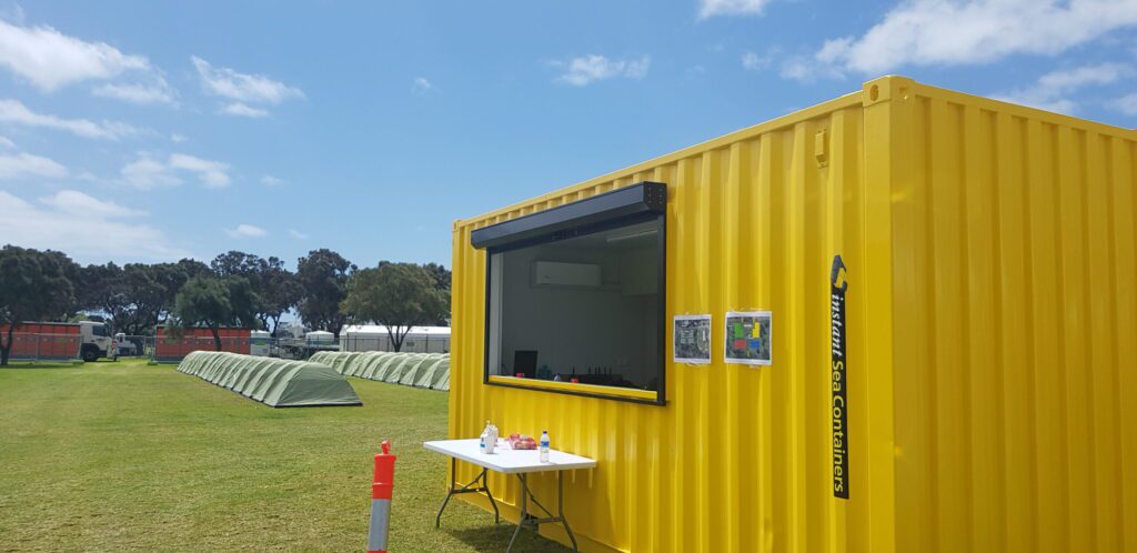 Bright yellow shipping container kitchen with an open service window on a grassy field, surrounded by tents and outdoor equipment.