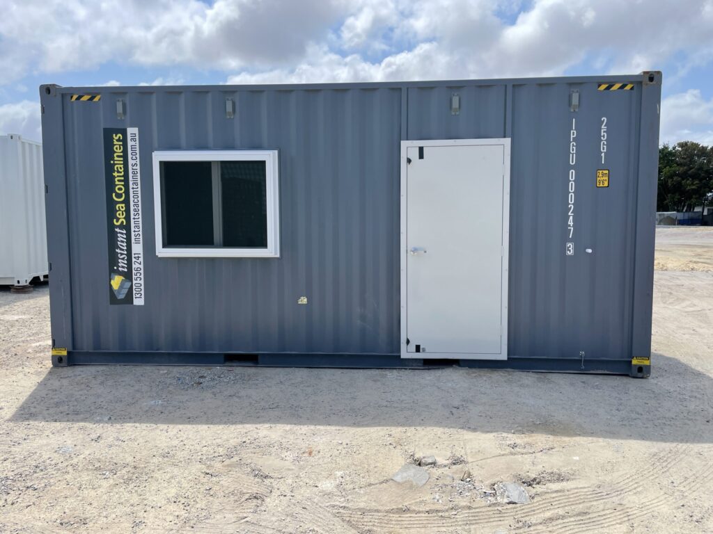 Gray shipping container office with a white door and single window, branded with Instant Sea Containers, placed on a dirt lot under a partly cloudy sky