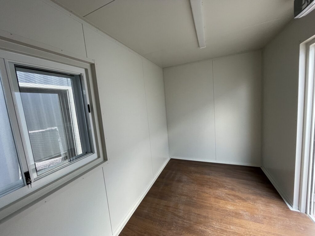 Interior of a small, empty white shipping container office with wood-look vinyl flooring and a small window.