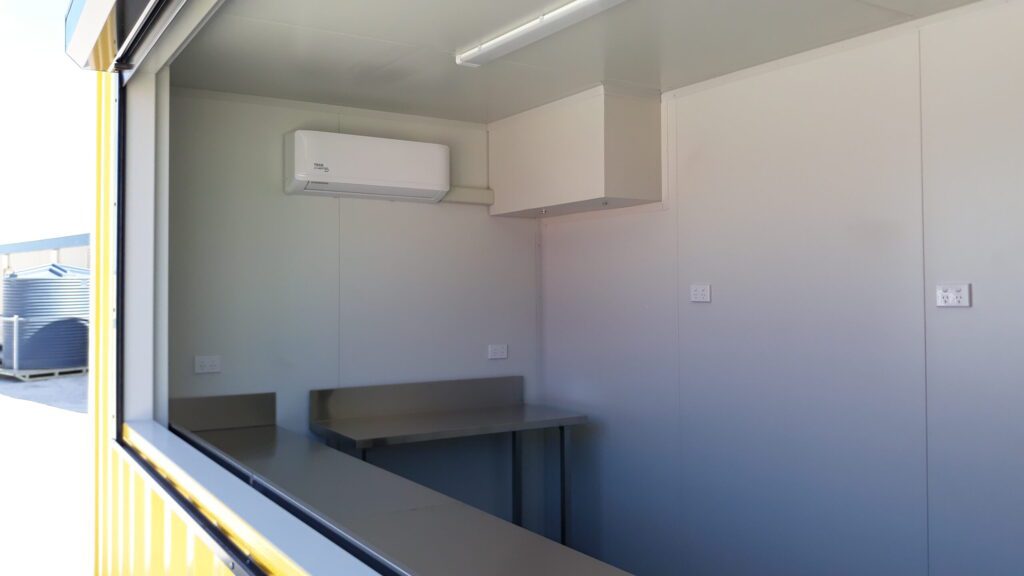 Interior view of a shipping container kitchen setup with stainless steel benches, wall-mounted air conditioner, and neutral gray walls.