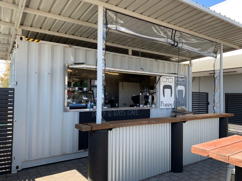 White shipping container kitchen setup with counter, transparent canopy, and “Poppy & Ray’s Cafe” signage, viewed from outside.