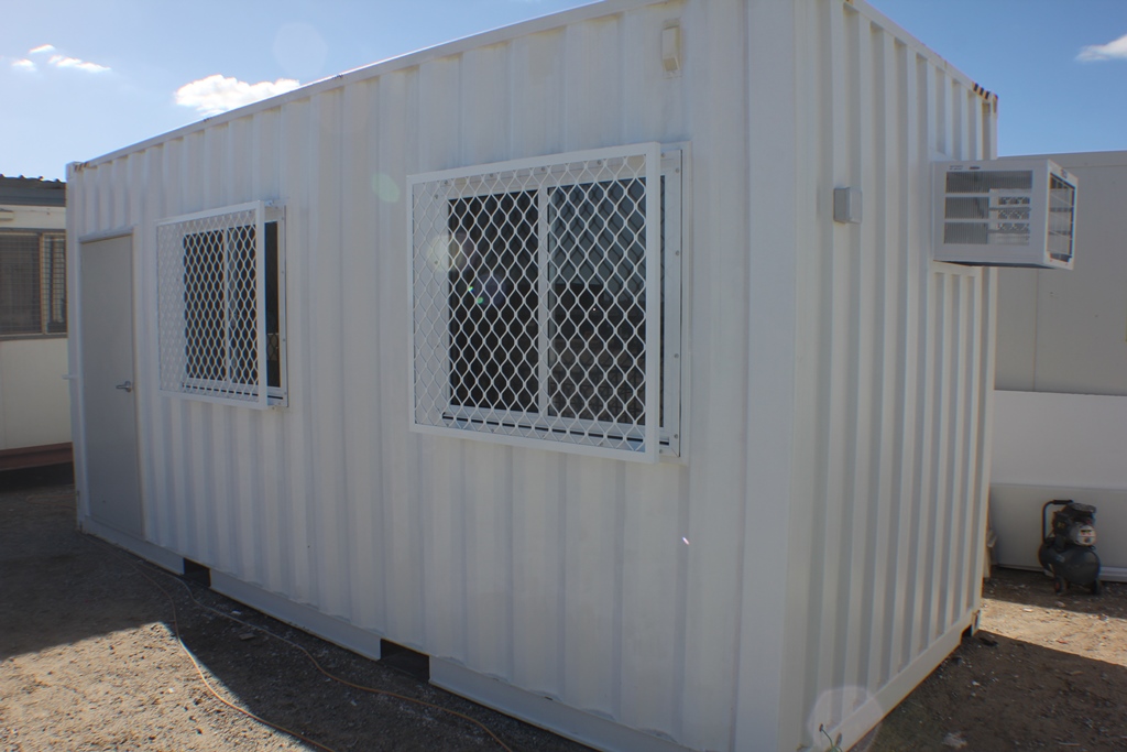 White shipping container office with two barred windows and an air conditioning unit, positioned outdoors on a sunny day.