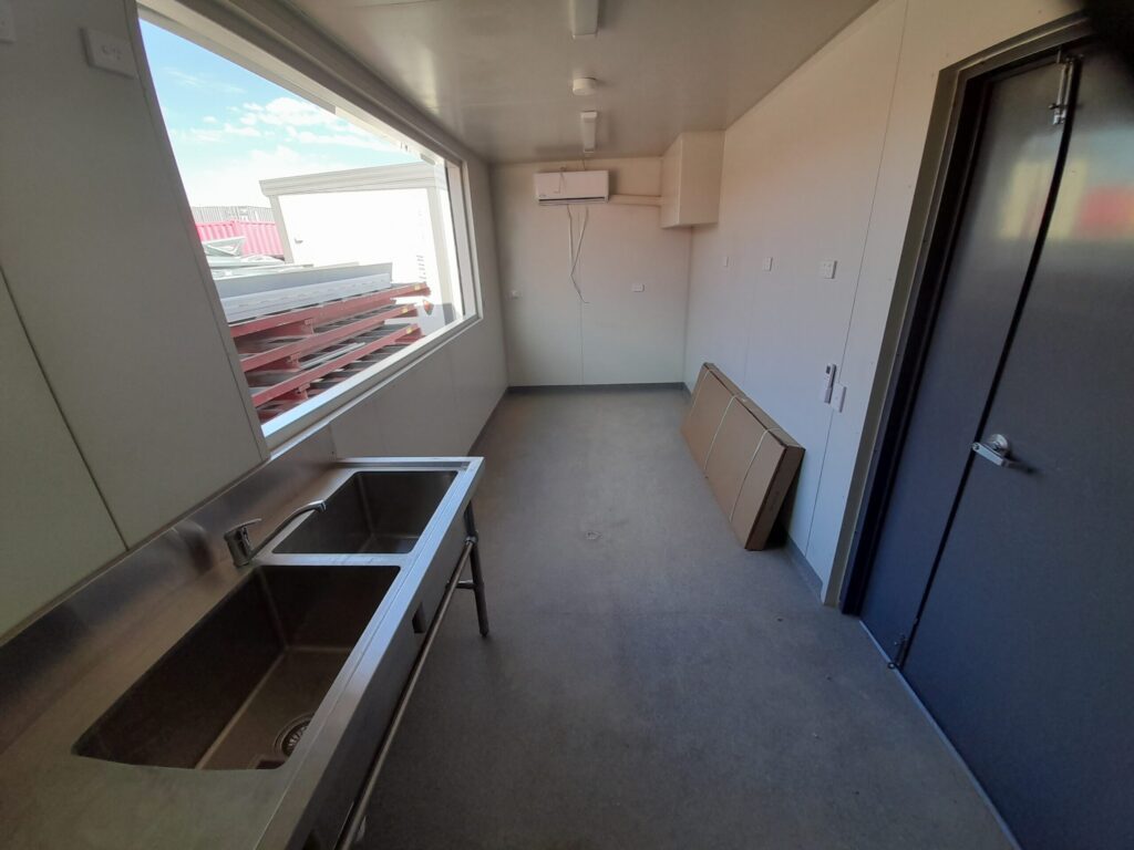 shipping container kitchen interior with stainless steel sink, bench space, large window, and storage items against the wall.